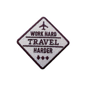 May include: A diamond-shaped embroidered patch with a white background and a dark burgundy border. The patch features an airplane icon and the text "WORK HARD TRAVEL HARDER" in burgundy. Three burgundy diamond shapes are at the bottom.