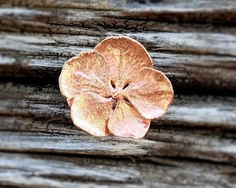 Op de afbeelding: Een koperkleurige bloemvormige broche met een ontwerp van vijf bloemblaadjes. De broche ligt op een stuk verweerd hout.