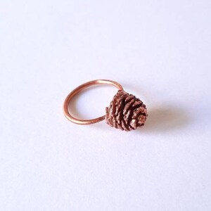 Può includere: Un anello in rame con un piccolo design di pigna dettagliato.