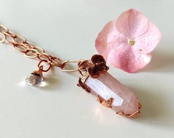 Op de afbeelding: Een ketting met een hanger van rozenkwartskristal, een koperen ketting en bloemmotieven. De hanger is een lange, puntige kristal met koperdraad eromheen gewikkeld. Het koperdraad is gevormd tot bladeren en bloemen.