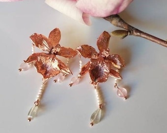 Op de afbeelding: Oorbellen van roségoud in de vorm van een orchidee met roze en witte kralen. De oorbellen hangen en hebben een delicaat bloemenmotief.