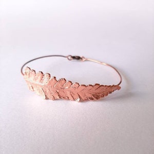 May include: A delicate copper bracelet featuring a fern leaf design. The bracelet is made of thin wire and has a simple, elegant look.