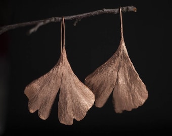 Può includere: Un paio di orecchini color rame a forma di foglie di ginkgo. Gli orecchini sono appesi a un ramo su uno sfondo nero.