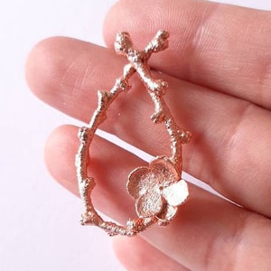 Pu&ograve; includere: Un ciondolo in oro rosa con un delicato design a ramo e un singolo fiore in basso.