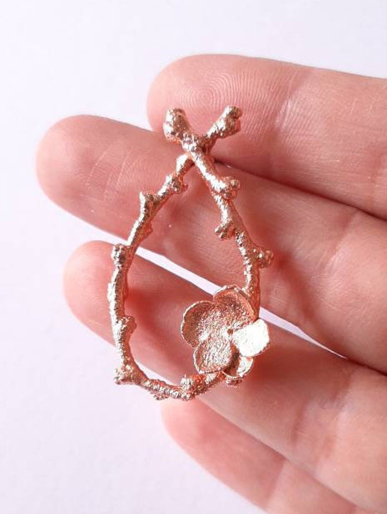 Pu&ograve; includere: Un ciondolo in oro rosa con un delicato design a ramo e un singolo fiore in basso.