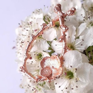 Pu&ograve; includere: Un ciondolo in metallo color oro rosa a forma di fiore con un gambo e foglie delicate. Il ciondolo &egrave; appoggiato su un gruppo di fiori bianchi.