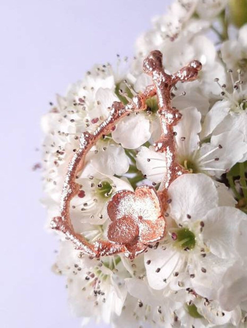 Pu&ograve; includere: Un ciondolo in metallo color oro rosa a forma di fiore con un gambo e foglie delicate. Il ciondolo &egrave; appoggiato su un gruppo di fiori bianchi.