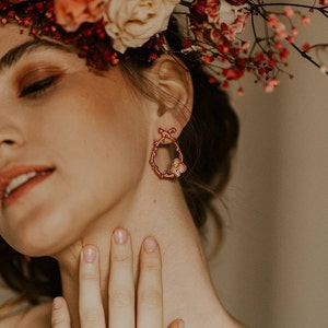 Pu&ograve; includere: Una donna indossa una corona di fiori con un paio di orecchini a cerchio in oro rosa con un design a farfalla. Indossa anche un anello al dito.