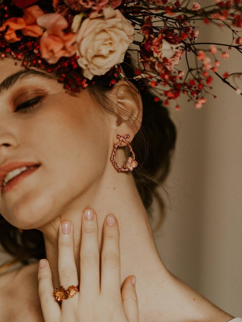 Pu&ograve; includere: Una donna indossa una corona di fiori con un paio di orecchini a cerchio in oro rosa con un design a farfalla. Indossa anche un anello al dito.