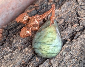 Op de afbeelding: Een druppelvormige labradoriet hanger met een glad, gepolijst oppervlak in iriserende groen- en grijstinten. De steen is bevestigd aan een textuurrijk, koperkleurig metalen takje met drie kleine, afgeronde, besachtige vormen.