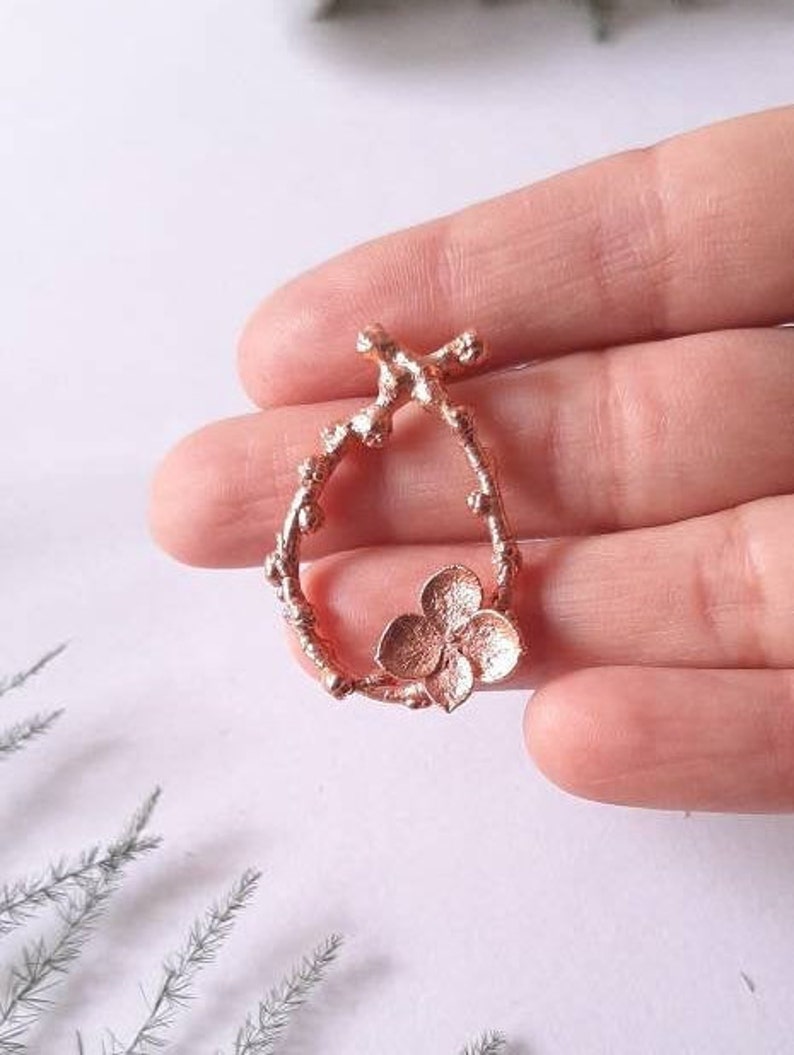Pu&ograve; includere: Un ciondolo in oro rosa con un delicato design floreale. Il ciondolo ha la forma di un ramo con un piccolo fiore al centro.