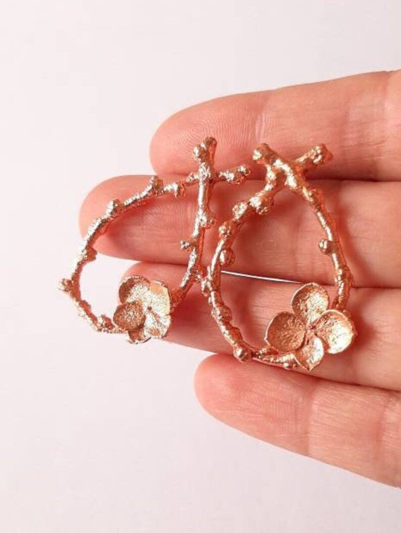 Pu&ograve; includere: Un paio di orecchini in oro rosa con un delicato design floreale. Gli orecchini sono in metallo e hanno una forma a ramo con piccoli accenti floreali.