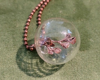 Op de afbeelding: Een koperen ketting met een glazen bol hanger die een delicate koperen tak met bladeren bevat.