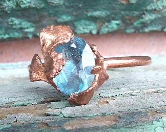 Op de afbeelding: Koperkleurige ring met een grote, lichtblauwe edelsteen. De edelsteen is onregelmatig van vorm en gezet in een getextureerde, organisch ogende zetting. De ring wordt getoond op een verweerd, teal-geverfd oppervlak.