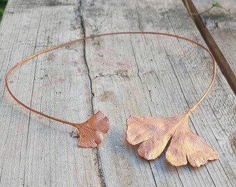 Op de afbeelding: Een koperkleurige ketting met ginkgo blad hangers. De ketting is gemaakt van een dunne draad die een cirkelvorm vormt, met twee ginkgo blad hangers aan de voorkant. De bladeren hebben gedetailleerde nerven en een getextureerd oppervlak.