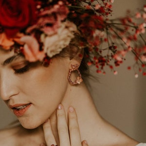 Pu&ograve; includere: Una donna con una corona di fiori con fiori rossi e rosa. Indossa un paio di orecchini a cerchio dorati con un design a farfalla e un anello dorato con un design a farfalla al dito.