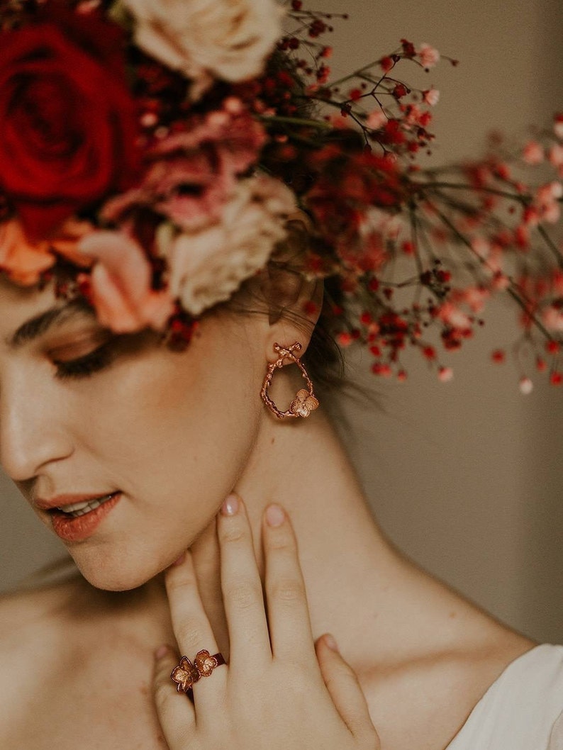 Pu&ograve; includere: Una donna con una corona di fiori con fiori rossi e rosa. Indossa un paio di orecchini a cerchio dorati con un design a farfalla e un anello dorato con un design a farfalla al dito.