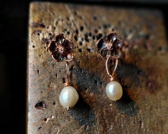Op de afbeelding: Een paar koperkleurige bloemvormige oorbellen met een witte parel die naar beneden hangt. De oorbellen zijn geplaatst op een houten oppervlak. Het bloemmotief is gedetailleerd en de parels zijn glad en rond.