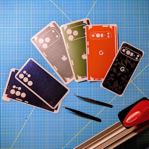 May include: A collection of phone skins in various colors, including blue, gray, green, orange, and black, with white accents. The skins feature cutouts for cameras and logos, along with a pair of black tweezers and a red and silver cutting tool on a blue grid surface.