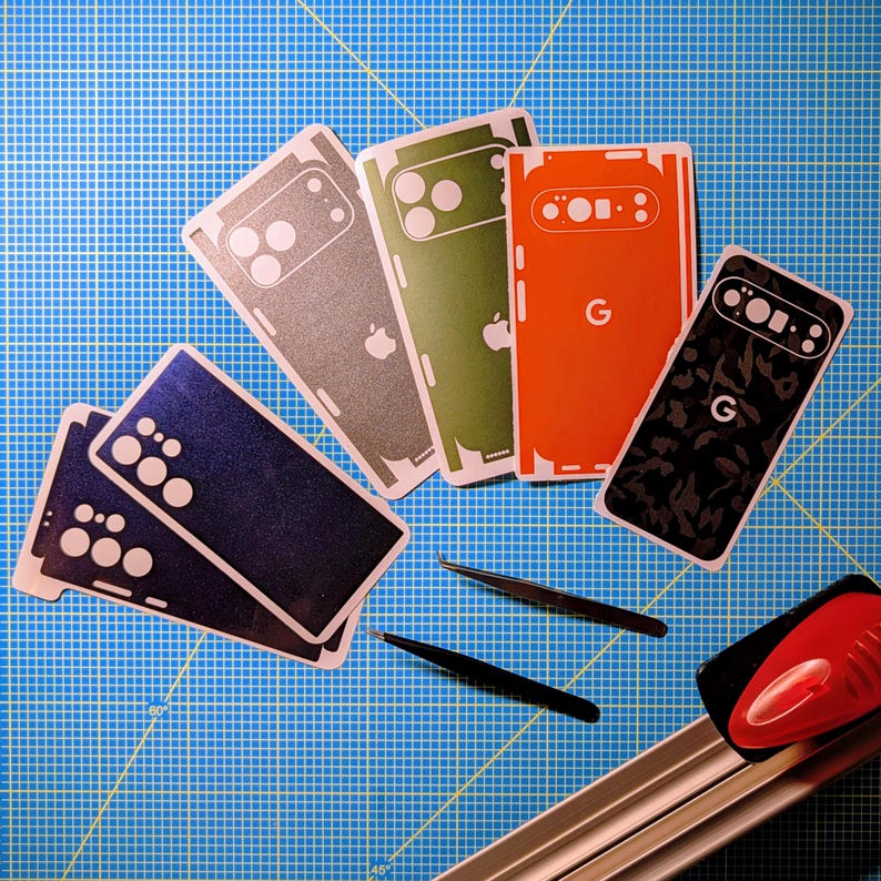 May include: A collection of phone skins in various colors, including blue, gray, green, orange, and black, with white accents. The skins feature cutouts for cameras and logos, along with a pair of black tweezers and a red and silver cutting tool on a blue grid surface.