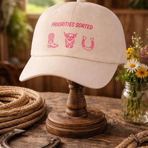 Può includere: Cappellino da baseball color crema con la scritta rosa "PRIORITIES SORTED" e grafiche rosa di uno stivale, un toro e un ferro di cavallo. Il cappellino è esposto su un supporto di legno, con un ferro di cavallo e una corda in primo piano.