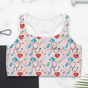 Puede incluir: Un sujetador deportivo rosa claro con interior blanco y un estampado de símbolos médicos, incluyendo estetoscopios, corazones, jeringas y gorros de enfermera. El sujetador está diseñado para actividades deportivas.