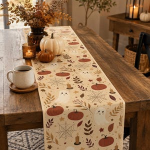 May include: A cream-colored table runner featuring a repeating pattern of white ghosts, orange pumpkins, spider webs, brown leaves, and small candles. The runner is laid out on a rustic wooden table alongside a white mug, a candle, and pumpkins.