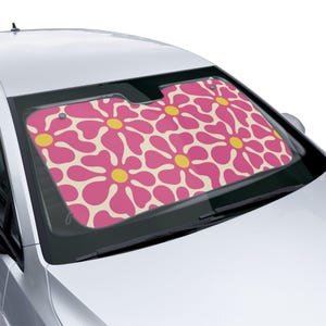 Op de afbeelding: Een zonnescherm voor de voorruit van een auto met een herhalend patroon van grote roze bloemen met gele harten op een crèmekleurige achtergrond. Het zonnescherm is geplaatst in de voorruit van een wit voertuig en bedekt het glas.
