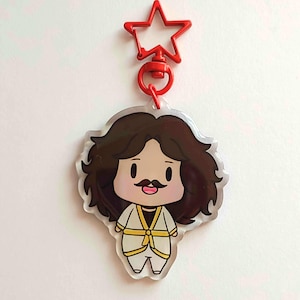 May include: A clear acrylic keychain featuring a cartoon character with brown hair, a mustache, and a white outfit with yellow accents. The keychain has a red star-shaped clasp.