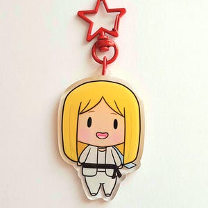 May include: A clear acrylic keychain featuring a cartoon illustration of a person with blonde hair, wearing a white uniform and a black belt. The keychain has a red star-shaped clip at the top.