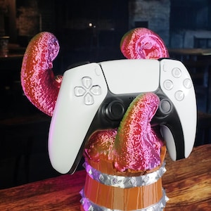 Puede incluir: Un mando blanco y negro de PlayStation 5 descansa sobre un soporte en forma de barril de madera con detalles plateados y tentáculos rojos, rosas y verdes. El mando tiene un diseño simétrico con botones y cruceta.