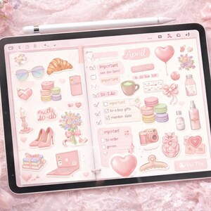 May include: A digital planner on a tablet, featuring a pink theme with stickers of a croissant, sunglasses, macarons, and hearts. The word "April" is visible, along with to-do lists and other decorative elements. A white stylus rests on top.