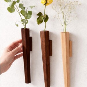 May include: Three wooden wall vases in varying shades of brown and natural wood. Each vase holds a different type of greenery and flowers. The vases are mounted on a white wall, creating a minimalist aesthetic.