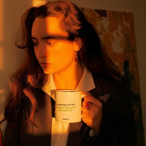 May include: White ceramic mug with black trim, featuring the text "I'm competing with YOU, I'm competing with YOUR PARENTS" in green. The mug is held by a person wearing a blazer and a white shirt. A red ring and earrings are also visible.