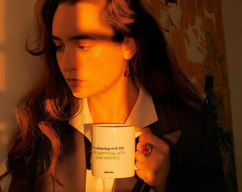 Puede incluir: Taza de cerámica blanca con borde negro, con el texto "I'm competing with YOU, I'm competing with YOUR PARENTS" en verde. La taza la sostiene una persona que lleva una blazer y una camisa blanca. También se ven un anillo y pendientes rojos.