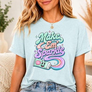 Op de afbeelding: Een lichtblauw t-shirt met een afbeelding van een lachende tandpastatube waar een kleurrijke, wervelende stroom tandpasta uit komt. De tekst "Make Em' Sparkle" staat in een retro lettertype, met "FRESH & CLEAN" op de tube gedrukt.