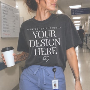 May include: A charcoal gray short-sleeve t-shirt featuring the white text GENTLEHEALTHSTUDIO, YOUR DESIGN HERE, and a small heart icon with an EKG line. The shirt is worn with light blue drawstring scrub pants and a visible ID badge clip.