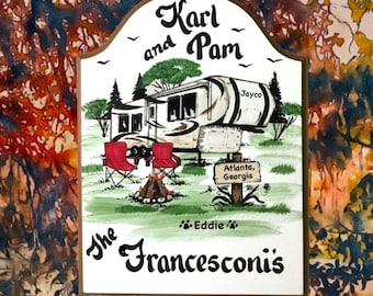 May include: A white wooden sign with a brown border, featuring a hand-painted illustration of a camper van, two red chairs, a campfire, and a sign that reads "Atlanta, Georgia". The text "Karl and Pam" is at the top of the sign, and the text "The Francesconis" is at the bottom. The text "Eddie" is written below the campfire.
