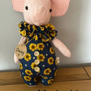 May include: A handmade pig doll with pink ears and a matching snout. The doll wears a navy blue jumpsuit adorned with yellow sunflowers and a ruffled collar. A tag reads "Daisy". The word "Marigold" is at the bottom.