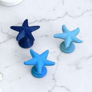 Op de afbeelding: Drie stervormige kastknoppen in blauwtinten, gerangschikt op een wit marmeren oppervlak. De knoppen zijn in verschillende blauwtinten, van diep marineblauw tot licht aqua. Zeeschelpen en een witte zeepbakje zijn ook zichtbaar.