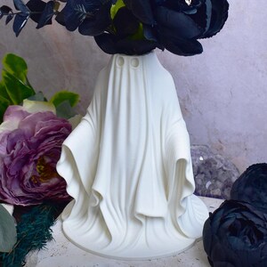 May include: A white ceramic ghost-shaped vase holding dark blue flowers and foliage. The vase has two eye holes and a draped design. Purple and green flowers are in the foreground. The background is a light purple and gray.