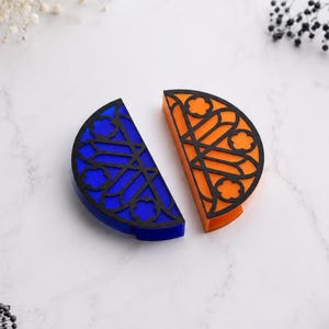Könnte beinhalten: Zwei halbkreisförmige Objekte mit einer schwarzen Gitterauflage in einem floralen und geometrischen Muster. Ein Teil hat eine leuchtend blaue Basis, während das andere eine leuchtend orangefarbene Basis hat. Beide Objekte sind halbkreisförmig.