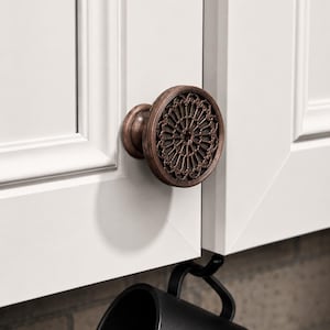 Puede incluir: Primer plano de un pomo de armario de bronce con un diseño decorativo circular de filigrana. El pomo está montado en una puerta de armario blanca. Una taza negra cuelga debajo del pomo.