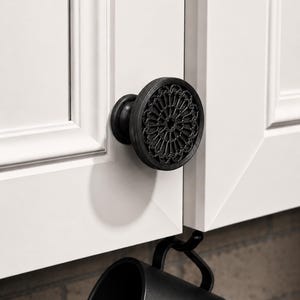 Puede incluir: Primer plano de un pomo de armario negro con un diseño circular decorativo. El pomo está montado en una puerta de armario blanca. Una taza negra cuelga debajo. El diseño del pomo presenta un patrón simétrico intrincado.