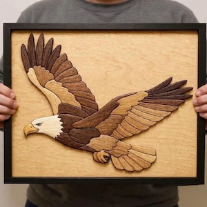 Puede incluir: Una obra de arte enmarcada en madera que representa un águila volando. El águila está hecha de diferentes tonos de madera marrón, con una cabeza blanca y un pico amarillo. La obra de arte está sobre un fondo marrón claro.