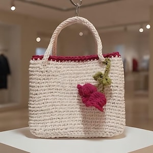 May include: A cream-colored, crocheted tote bag with a burgundy trim and handle. A pink and green floral embellishment is attached to the front. The bag is displayed on a white pedestal.