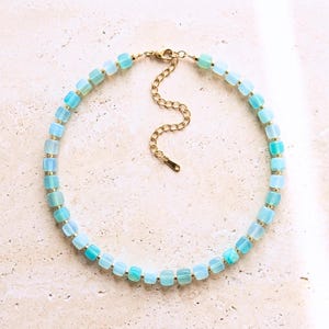 Può includere: Una collana con perline cubiche semitraslucide color azzurro alternate a piccoli distanziatori in metallo dorato. La collana include una chiusura a moschettone in metallo dorato e una catenella di estensione regolabile con una piccola targhetta.