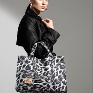 May include: A rectangular tote bag featuring a black and white leopard print pattern. The bag has two top handles and a rectangular tan leather-coloured patch on the lower left side with the text Q.Nary printed in black ink on the surface.