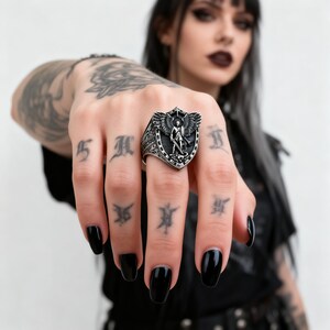 May include: A silver ring with a shield-shaped face featuring an angel with outstretched wings and a sword. The ring has a detailed, gothic design. The person in the image has black nail polish.