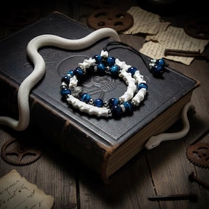 Puede incluir: Un collar artesanal con cuentas de ojo de tigre azul y cuentas blancas en forma de hueso. El collar se exhibe sobre un libro oscuro con una figura de serpiente blanca. El fondo incluye engranajes antiguos y papel envejecido.